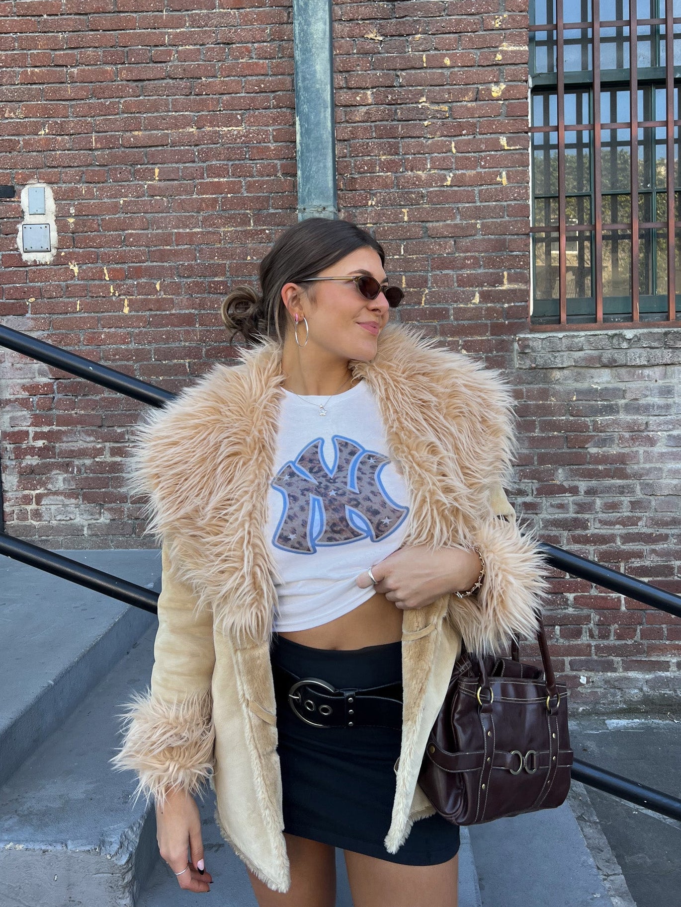 Woman wearing a fluffy beige jacket and white graphic t-shirt in front of a brick building.